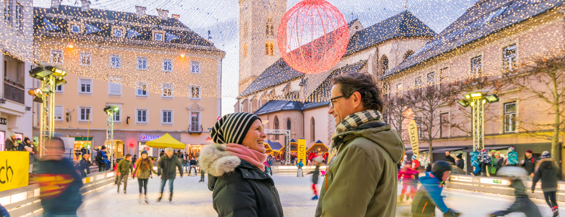 Zwei Personen auf einem festlich beleuchteten Platz mit Eislaufbahn in einer Kärntner Altstadt