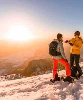 Zwei Personen mit Schneeschuhen auf einem verschneiten Berggipfel bei Sonnenuntergang