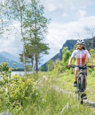 Radfahrerin auf einem Radweg am Flussufer vor Kärntner Bergkulisse