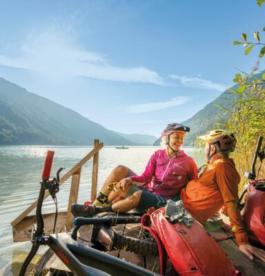 Zwei Radreisende sitzen an einem Seeufer vor Bergkulisse