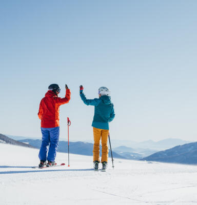 Zwei Skifahrende stehen auf einer verschneiten Piste und begrüßen sich mit erhobenen Händen. 