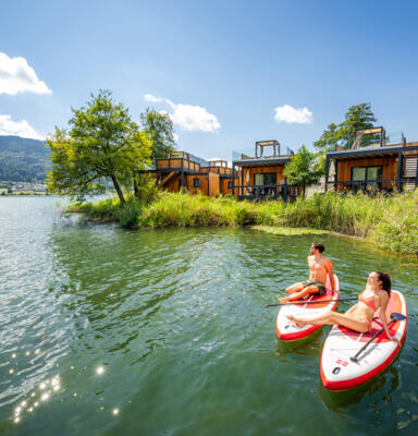 34_Kärnten Werbung GmbH_Michael Stabentheiner Zwei Personen sitzen auf Stand-up-Paddle-Boards vor Ferienanlagen am See