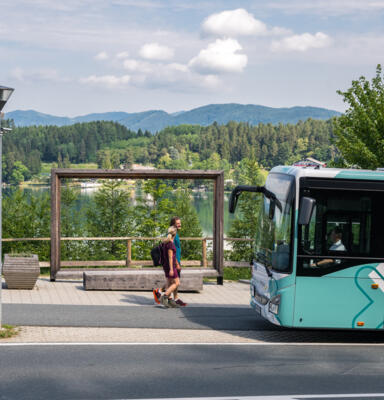 Zwei Personen vor einem Regionalbus an einer Haltestelle vor Seen- und Berglandschaft 