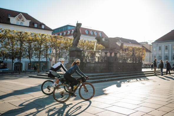 11_Meridiem Trail_Martin Hofmann Zwei Radfahrende auf einem Platz in einer Kärntner Innenstadt